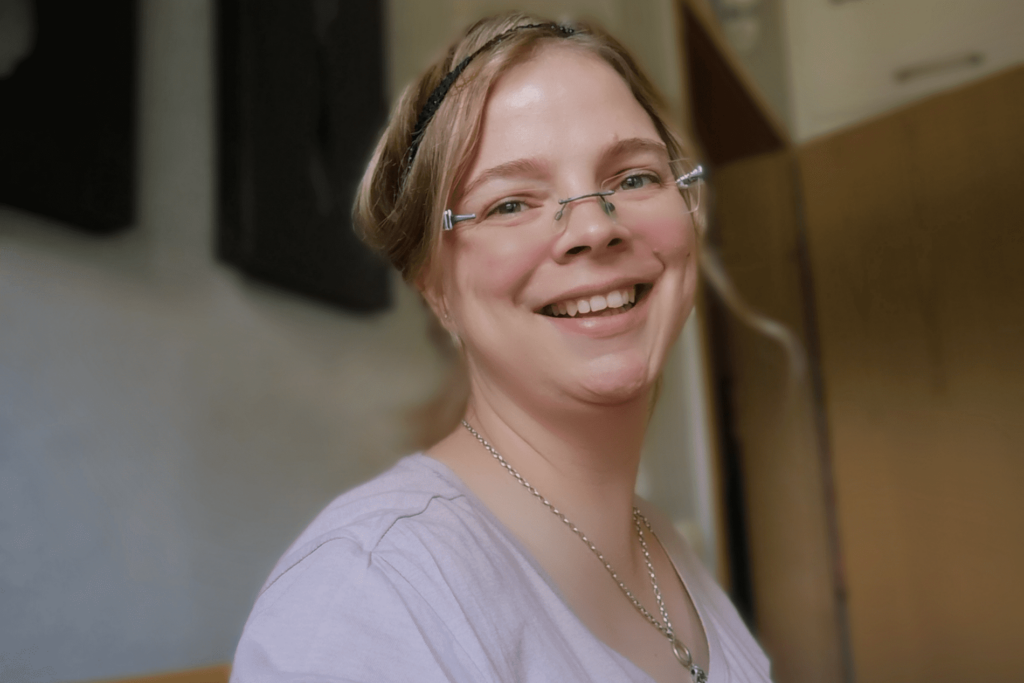 Lachendes Porträt von Maggie in heller Kleidung mit Brille und geflochtenem Haarband, aufgenommen in Innenraum
