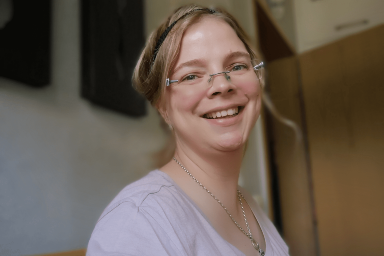 Lachendes Porträt von Maggie in heller Kleidung mit Brille und geflochtenem Haarband, aufgenommen in Innenraum